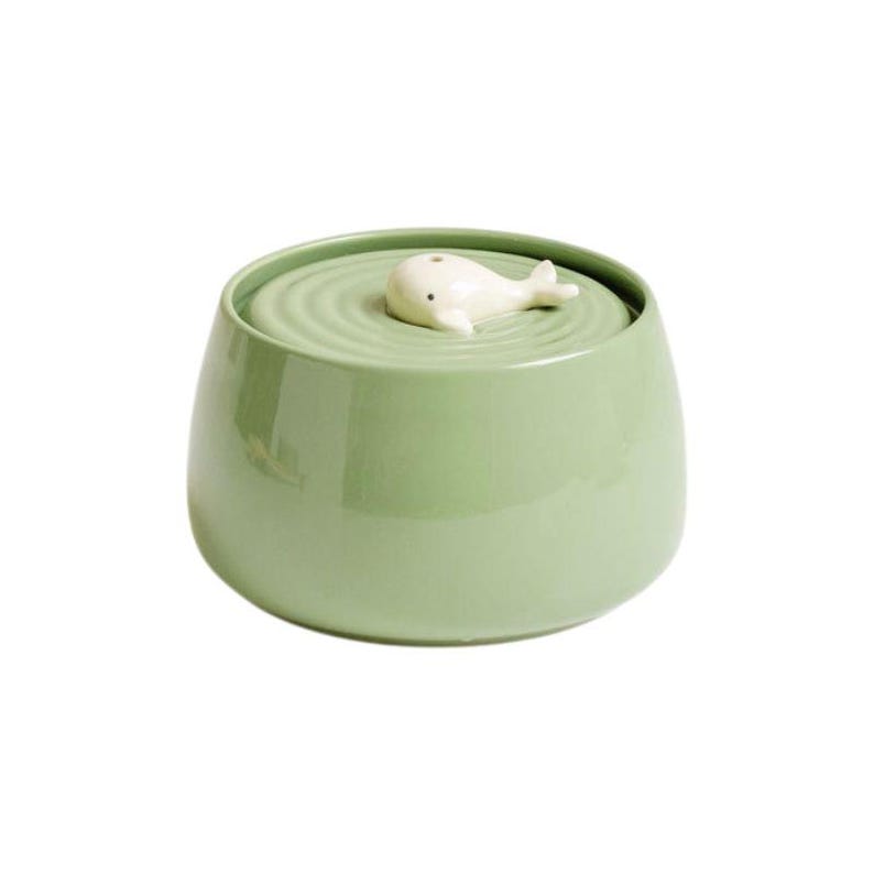 May include: A light green ceramic jar with a lid featuring a white whale figurine. The jar has a rounded shape and a glossy finish. The lid has concentric circle detailing. The whale figurine is small and white.