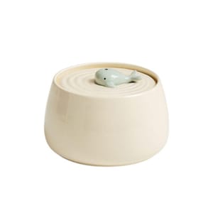 May include: Cream-colored ceramic jar with a lid featuring a light blue whale figurine. The lid has concentric circle detailing. The jar has a rounded shape and a smooth, glossy finish. A decorative storage container.