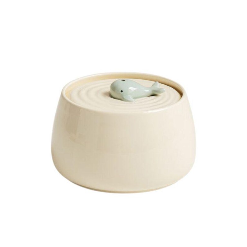 May include: Cream-colored ceramic jar with a lid featuring a light blue whale figurine. The lid has concentric circle detailing. The jar has a rounded shape and a smooth, glossy finish. A decorative storage container.