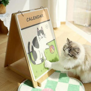 Op de afbeelding: Een houten ezelkalender met een cartoonkattenillustratie. Het woord "CALENDAR" staat bovenaan. Een pluizige kat interageert met de kalender. De kalender staat op een groen-wit geruite mat.