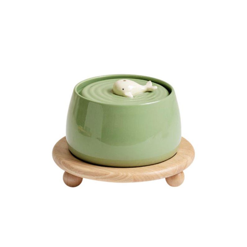 May include: A green ceramic bowl with a whale-shaped lid, resting on a round wooden base with three small, rounded feet. The bowl has a smooth, glossy finish, and the whale is white.