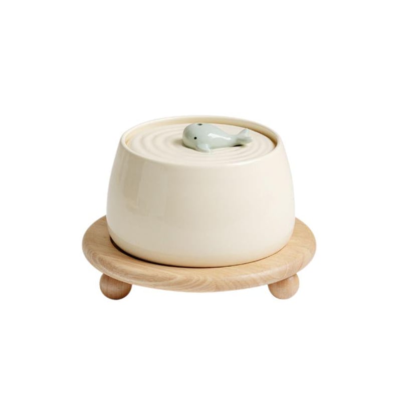 May include: A cream-colored ceramic container with a whale-shaped lid, set on a round wooden stand with three small feet. The whale is light blue. The container has a smooth, glossy finish.