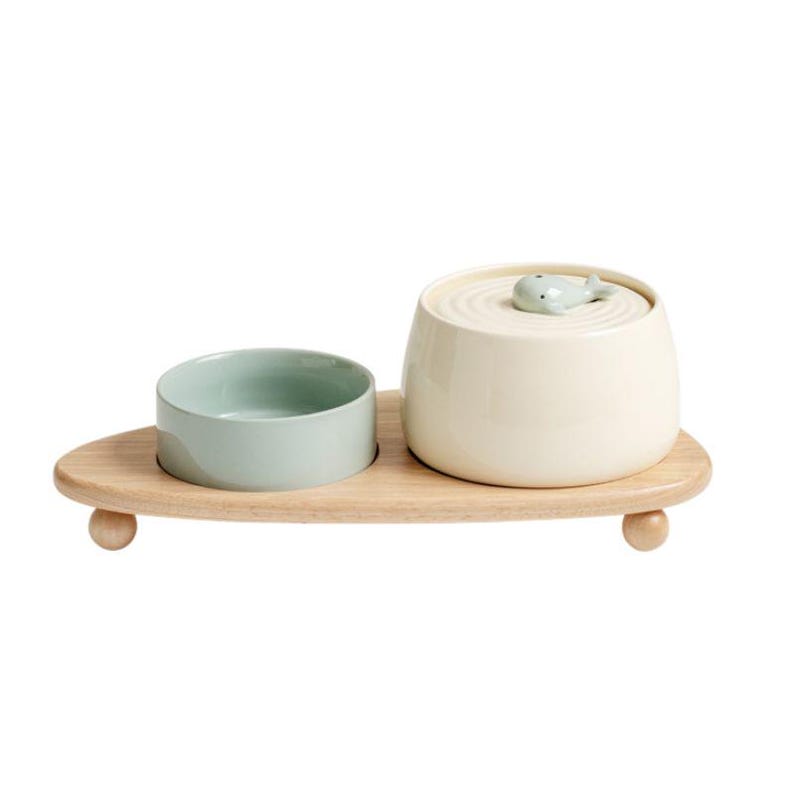 May include: A light wood serving tray with a small, round, light green bowl and a cream-colored container with a lid. The lid has a small, light green whale-shaped handle. The tray has small, round feet.