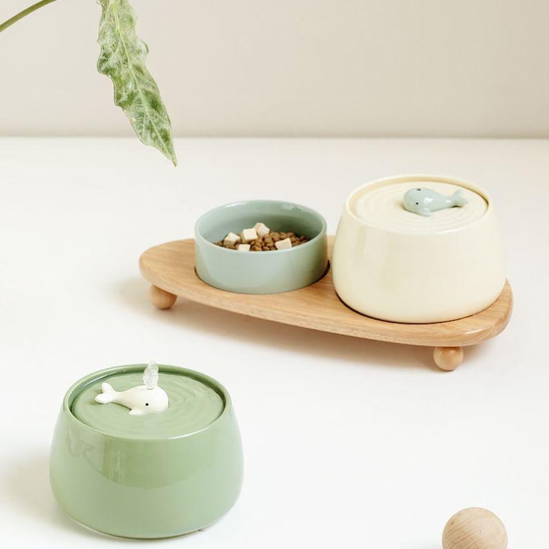 May include: A wooden pet food and water bowl set. The set includes a light green bowl with food, a cream-colored bowl with a whale-shaped lid, and a green bowl with a whale-shaped fountain. The bowls sit on a wooden tray.