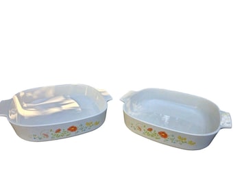 Op de afbeelding: Twee rechthoekige witte keramische ovenschalen met geïntegreerde handgrepen aan de zijkanten. Elke schaal heeft een bloemmotief aan de buitenkant, met oranje klaprozen, gele bloesems en groen blad op een glad, glanzend wit oppervlak.