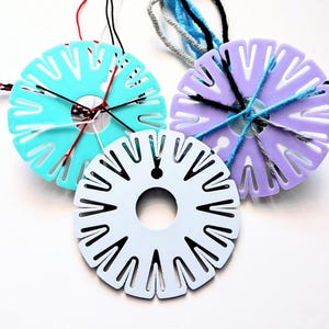 Puede incluir: Tres herramientas de tejido de plástico en color turquesa, lavanda y blanco, cada una con un agujero central y ranuras radiales. Las herramientas turquesa y lavanda tienen hilo enhebrado, con hebras rojas, negras, azules y grises visibles. Estas herramientas se utilizan para hacer pulseras de la amistad.