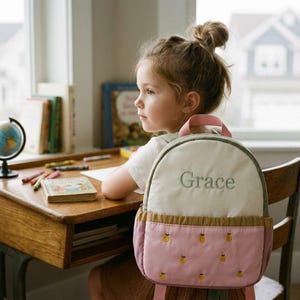 Puede incluir: Una mochila infantil con el nombre "Grace" bordado en la parte delantera. La mochila tiene una parte superior color crema, una parte inferior rosa con diseños de frutas amarillas y correas rosas. El niño está sentado en un escritorio de madera con libros y crayones.