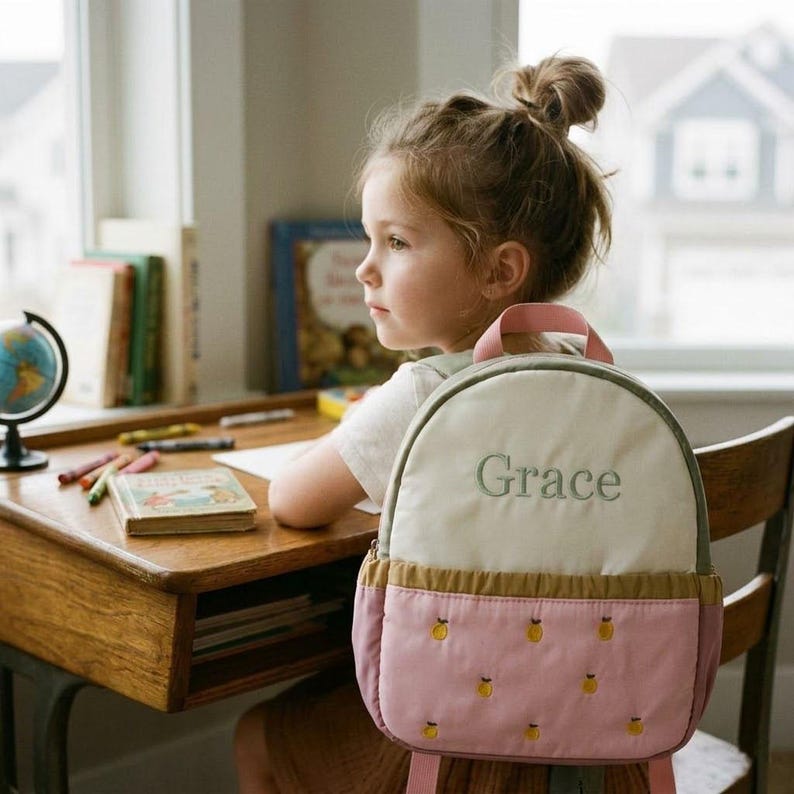 May include: A child's backpack with the name "Grace" embroidered on the front. The backpack has a cream-colored top, a pink bottom with yellow fruit designs, and pink straps. The child is sitting at a wooden desk with books and crayons.