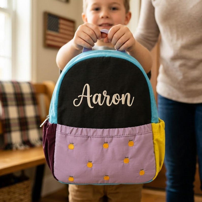 May include: A multi-colored backpack with the name "Aaron" in white script. The backpack has a black top, light blue trim, a purple pocket with embroidered orange fruit, and yellow side panels. The backpack is held by a child.