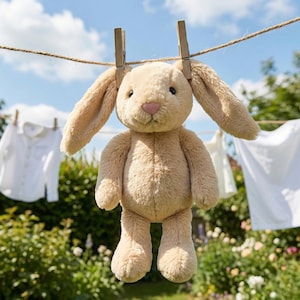 May include: A plush, tan-colored bunny toy with long floppy ears is suspended from a clothesline by wooden clothespins. The bunny has a pink nose and is hanging in front of a sunny, green garden with white shirts hanging on a clothesline.