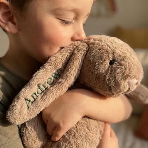 Puede incluir: Un peluche de conejo marrón claro con orejas largas y caídas. El nombre "Andrew" está bordado en verde en una oreja. El conejo está siendo abrazado, con la cara del niño parcialmente visible.