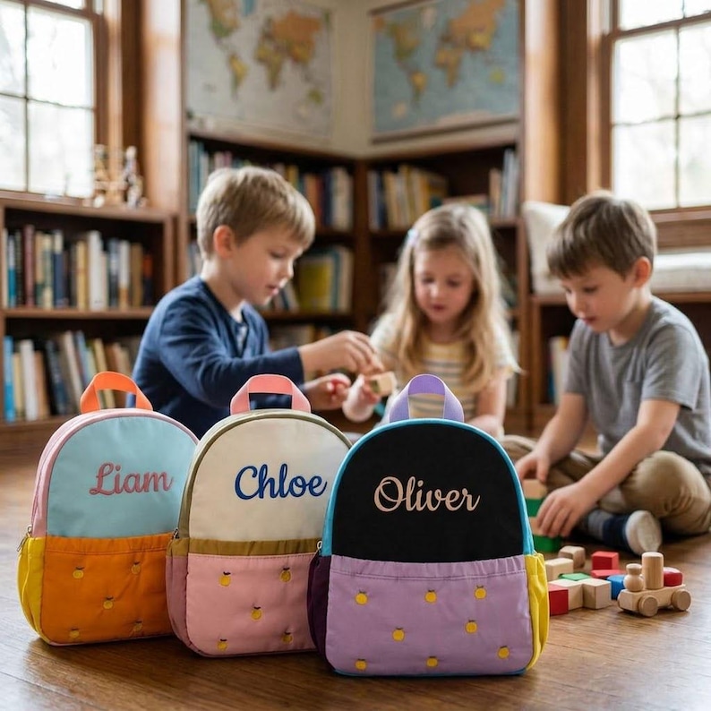 May include: Three personalized backpacks in the foreground, each with a different color scheme and name embroidered on the front: Liam, Chloe, and Oliver. The backpacks have a pineapple pattern on the bottom and are in front of children playing with blocks.