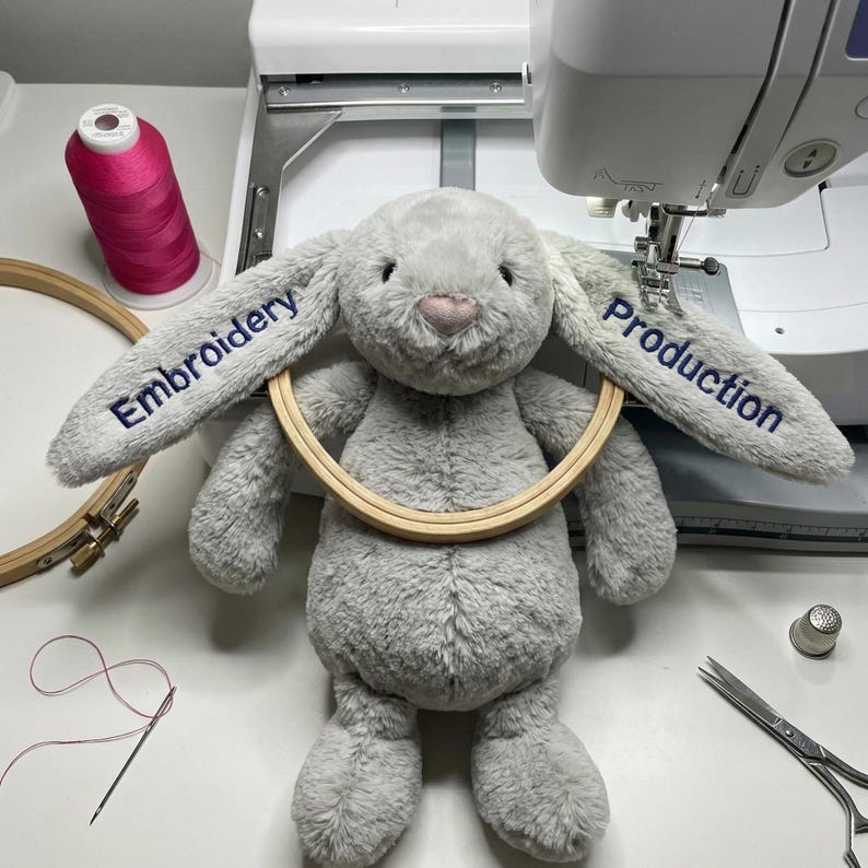 May include: A plush gray bunny with the words "Embroidery" and "Production" embroidered on its ears. The bunny is holding a wooden embroidery hoop. A spool of pink thread, a needle, and scissors are also visible.