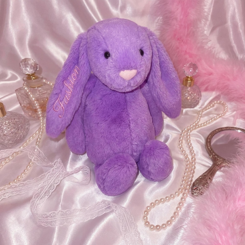 May include: A plush, lavender-colored bunny with long, floppy ears. The word "Fashion" is embroidered on one ear. The bunny has a pink nose and is surrounded by decorative items, including a pearl necklace and a vintage-style mirror.