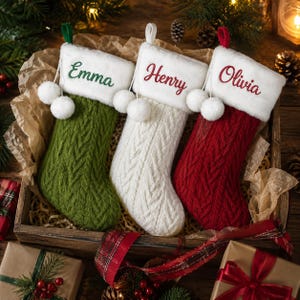 Peut inclure: Trois chaussettes de Noël en tricot torsadé vert, blanc et rouge, chacune dotée d'un revers en peluche blanche avec deux pompons blancs. Les revers sont brodés avec les prénoms Emma en vert, Henry en rouge et Olivia en rouge.