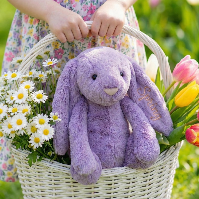 May include: A plush, lavender bunny with the word "Spring" embroidered on its ear sits in a white wicker basket filled with daisies and tulips. The bunny has long floppy ears and a soft, fuzzy texture.