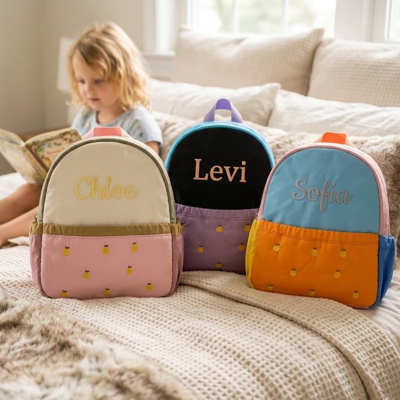 May include: Three colorful children's backpacks with embroidered names: Chloe, Levi, and Sofia. The backpacks feature a variety of colors, including pink, cream, blue, orange, and purple. Each backpack has a pineapple pattern on the bottom.