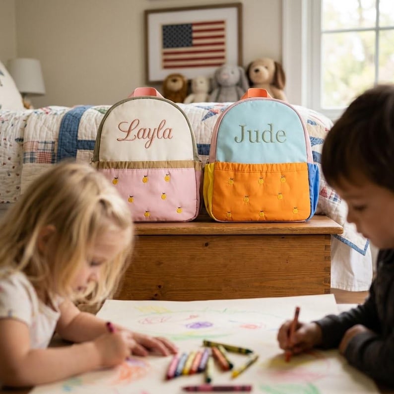 May include: Two personalized children's backpacks. One is cream and pink with the name "Layla" embroidered on it. The other is blue and orange with the name "Jude" embroidered. Both have a pineapple pattern on the front pocket.