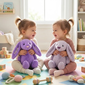 May include: Two plush bunny rabbits in shades of purple and lavender are held by children. The bunnies have long ears and are embroidered with names and numbers. The children are sitting on a colorful play mat.