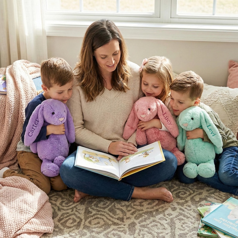 May include: A woman reads a book with three children, each holding a plush bunny. The bunnies are purple, pink, and teal, with names embroidered on them. The scene takes place indoors with a window in the background.