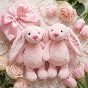 May include: Two plush pink bunny rabbits with long ears and pink heart-shaped noses. One ear reads "Forever" and the other reads "Gift". A pink gift box with a bow, pearls, and pink flowers surround the bunnies.