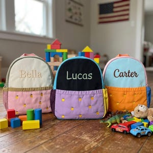 May include: Three personalized children's backpacks in pastel colors. Each backpack has a different name embroidered on the front: "Bella", "Lucas", and "Carter". The backpacks feature a front pocket with a yellow fruit pattern.