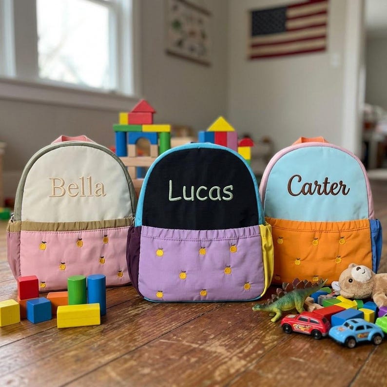 May include: Three personalized children's backpacks in pastel colors. Each backpack has a different name embroidered on the front: "Bella", "Lucas", and "Carter". The backpacks feature a front pocket with a yellow fruit pattern.