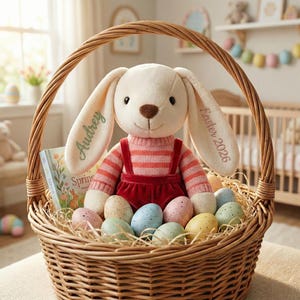 May include: A woven wicker basket filled with Easter decorations. A plush bunny wearing a pink striped sweater and red overalls sits in the basket. The bunny's ears are embroidered with the name "Audrey" and "Easter 2026". The basket also contains speckled Easter eggs and a children's book.