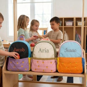 May include: Three personalized backpacks in black, cream, and light blue, each with a different name embroidered on the front: Jacob, Aria, and Isaac. The backpacks have colorful accents and a pineapple pattern on the front pocket.