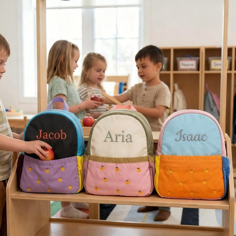 May include: Three personalized backpacks in black, cream, and light blue, each with a different name embroidered on the front: Jacob, Aria, and Isaac. The backpacks have colorful accents and a pineapple pattern on the front pocket.