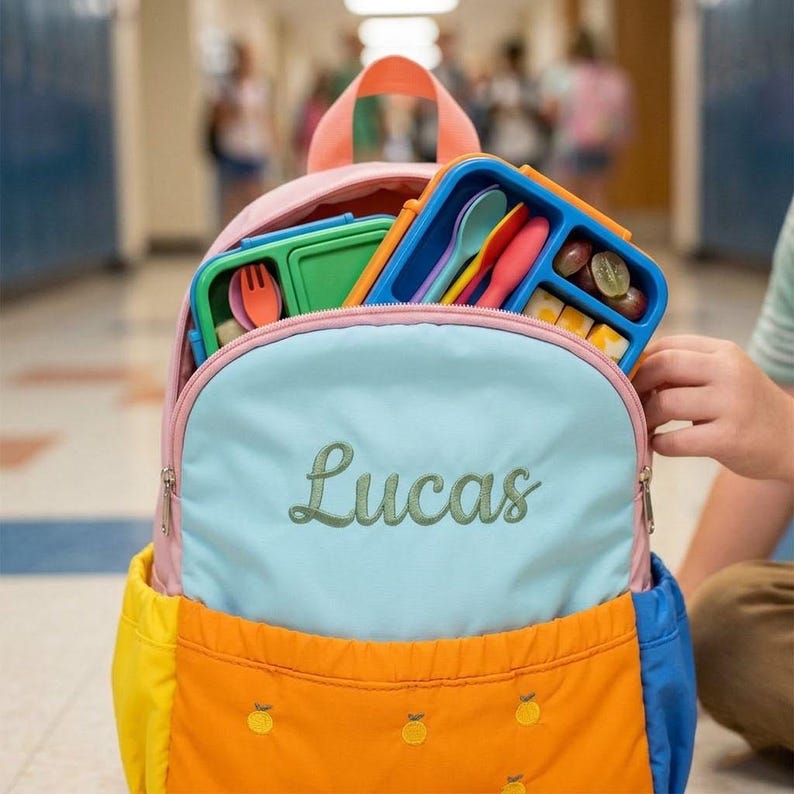 May include: A colorful backpack with the name "Lucas" embroidered on the front. The backpack is open, revealing a blue and green lunchbox with utensils and food. The backpack has a pink top, a light blue front, and a yellow and blue bottom.