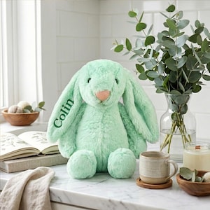 May include: A mint green plush bunny with long floppy ears and a pink nose. The name "Colin" is embroidered on one ear in green. The bunny is sitting on a white countertop next to a mug, a candle, and a vase of eucalyptus.