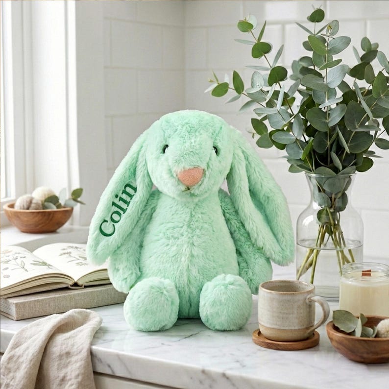 May include: A mint green plush bunny with long floppy ears and a pink nose. The name "Colin" is embroidered on one ear in green. The bunny is sitting on a white countertop next to a mug, a candle, and a vase of eucalyptus.