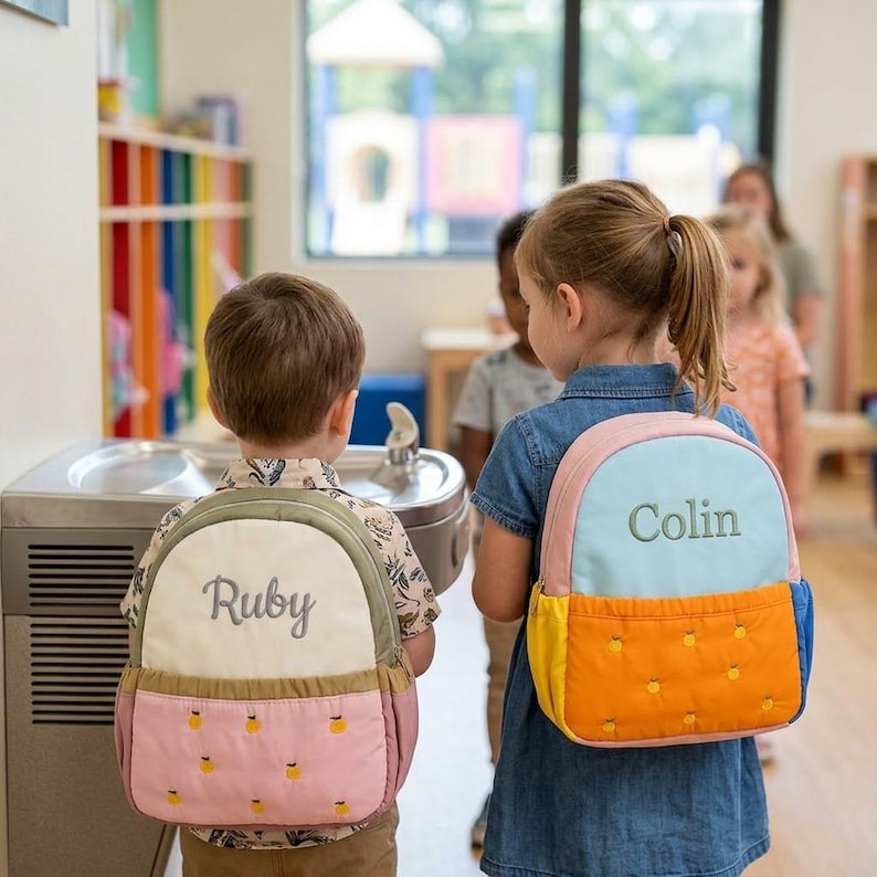 May include: Two children wearing personalized backpacks. One backpack is light blue, pink, and yellow with the name "Colin" embroidered on it. The other backpack is pink, green, and cream with the name "Ruby" embroidered on it. Both backpacks have a quilted design.