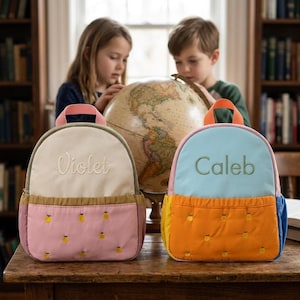 May include: Two personalized children's backpacks. One is cream, pink, and brown with the name "Violet" embroidered on the front. The other is blue, orange, and yellow with the name "Caleb" embroidered on the front. Both have a quilted design.