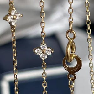 May include: Close-up of a gold necklace with a delicate chain. The necklace features four-petal flower pendants with clear gemstones. The clasp is marked with "K18 HIKAGE".