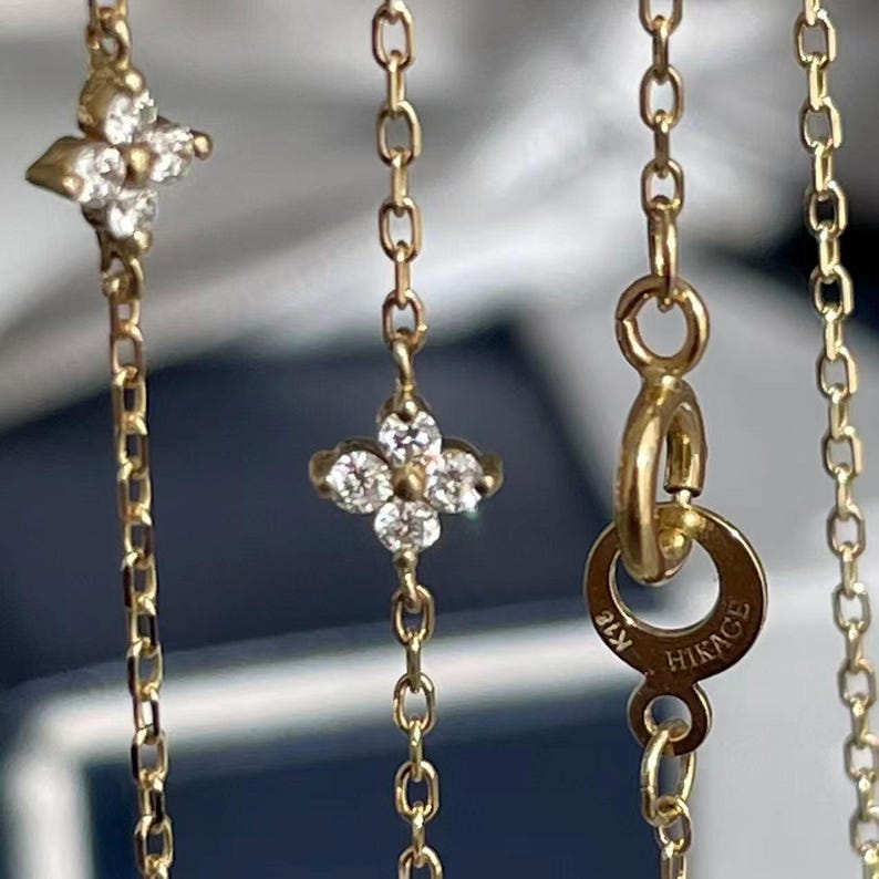 May include: Close-up of a gold necklace with a delicate chain. The necklace features four-petal flower pendants with clear gemstones. The clasp is marked with "K18 HIKAGE".