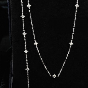 May include: A delicate silver necklace featuring small, sparkling, four-petal flower-shaped pendants. The necklace has a Y-shaped design, with a longer strand of flowers hanging down. The jewellery is displayed against a black background.