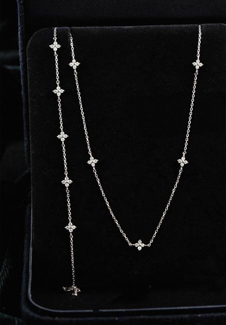 May include: A delicate silver necklace featuring small, sparkling, four-petal flower-shaped pendants. The necklace has a Y-shaped design, with a longer strand of flowers hanging down. The jewellery is displayed against a black background.
