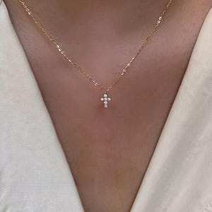 May include: A gold-colored necklace with a small cross pendant. The cross is adorned with multiple small, clear gemstones. The necklace is displayed against a neutral background.