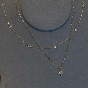 May include: A layered gold necklace with delicate chains. The top chain features small, evenly spaced diamond-like accents. The second chain has a cross pendant with a cluster of small stones. The necklace is displayed on a grey surface.