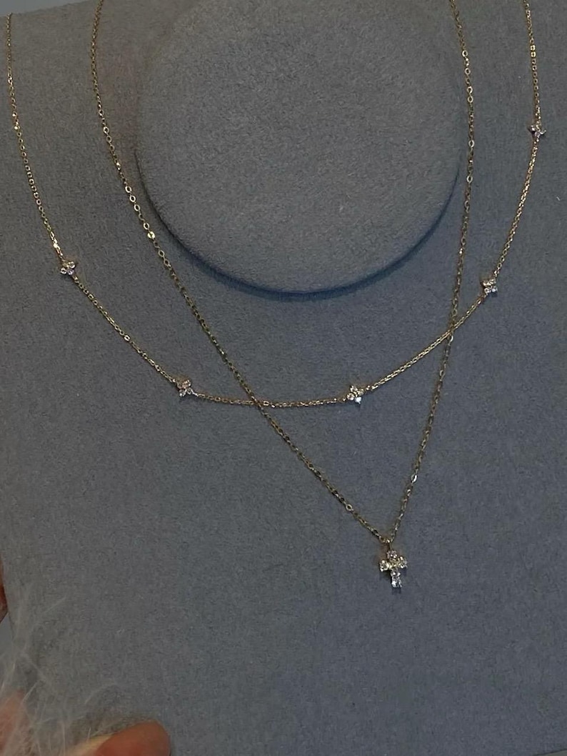 May include: A layered gold necklace with delicate chains. The top chain features small, evenly spaced diamond-like accents. The second chain has a cross pendant with a cluster of small stones. The necklace is displayed on a grey surface.