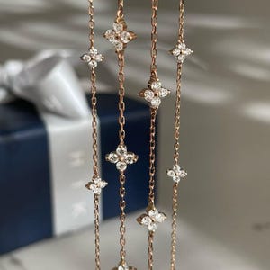 May include: Rose gold chain jewellery featuring multiple four-leaf clover pendants, each adorned with sparkling clear gemstones. The delicate chains hang vertically, creating a graceful and elegant display. The background is blurred, focusing attention on the jewellery.