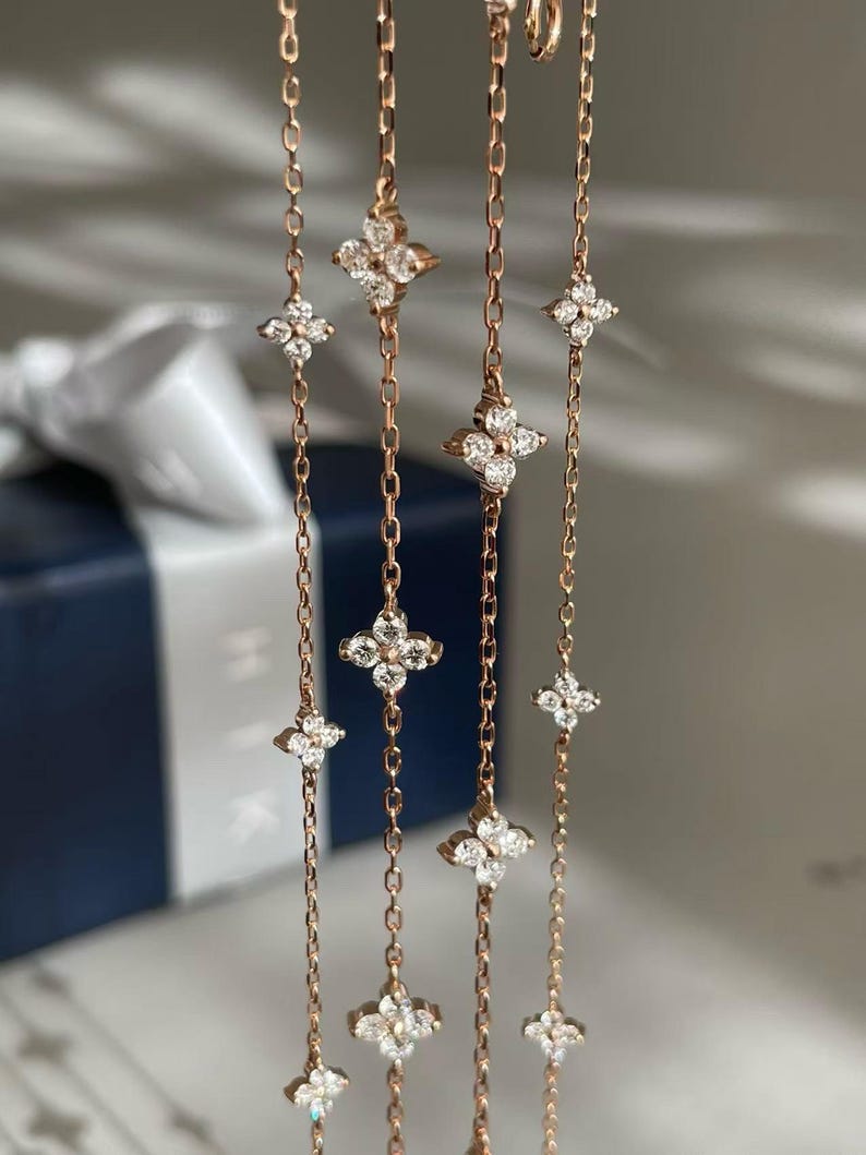 May include: Rose gold chain jewellery featuring multiple four-leaf clover pendants, each adorned with sparkling clear gemstones. The delicate chains hang vertically, creating a graceful and elegant display. The background is blurred, focusing attention on the jewellery.