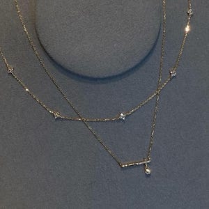 May include: Two delicate gold necklaces with small, sparkling accents. The top necklace features evenly spaced, tiny floral designs. The bottom necklace has a horizontal bar with a dangling charm. The necklaces are displayed on a grey surface.