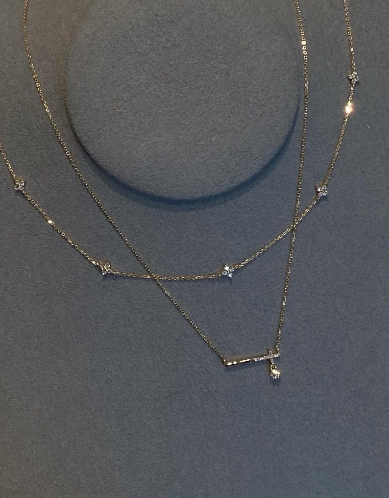 May include: Two delicate gold necklaces with small, sparkling accents. The top necklace features evenly spaced, tiny floral designs. The bottom necklace has a horizontal bar with a dangling charm. The necklaces are displayed on a grey surface.