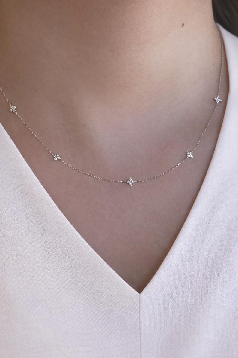 May include: A fine silver necklace with small, flower-shaped pendants. The necklace is displayed against a white top. The design is simple and elegant. A refined accessory for everyday wear or special occasions.