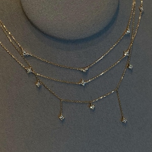 May include: A delicate gold necklace with three strands, each adorned with small, sparkling gemstones. The necklace features a layered design, with the gemstones evenly spaced along the chains. The necklace is displayed against a grey background.