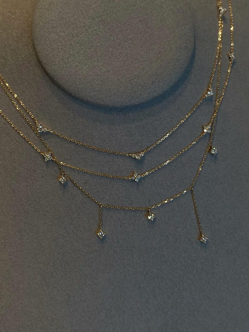 May include: A delicate gold necklace with three strands, each adorned with small, sparkling gemstones. The necklace features a layered design, with the gemstones evenly spaced along the chains. The necklace is displayed against a grey background.