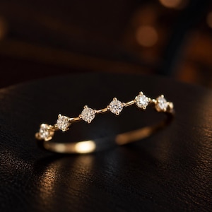 Puede incluir: Un anillo de banda fina de oro con una serie de seis pequeñas piedras redondas y transparentes talladas, engastadas individualmente con garras a lo largo del arco superior. El metal tiene un acabado en oro amarillo pulido y un diseño lineal.